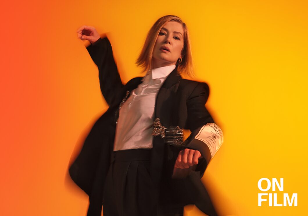 Actor Rosamund Pike in Inter Alia. She is wearing a suit and standing in front of a bright orange background.