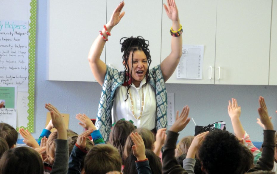Storytelling teaching artist leads young students in physical movements in front of classroom. Everyone's arms are up in the air.