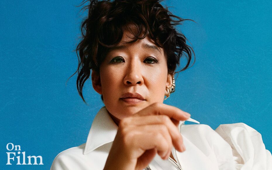 Photograph of Sandra Oh wearing a white blouse with her hair tied up against a blue background. Her hand partially covers her body.