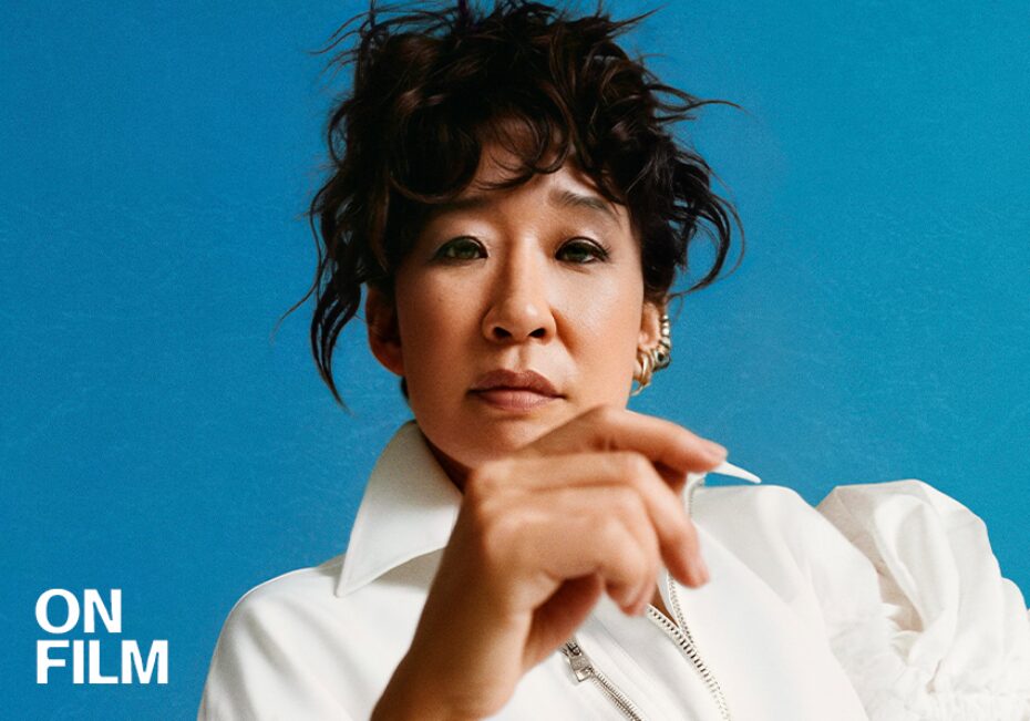 Photograph of Sandra Oh wearing a white blouse with her hair tied up against a blue background. Her hand partially covers her body.