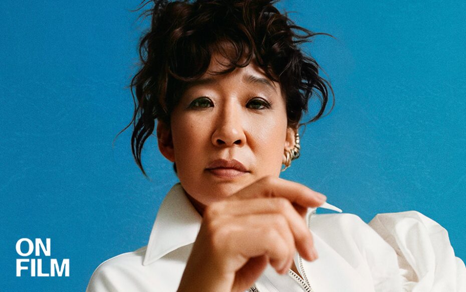 Photograph of Sandra Oh wearing a white blouse with her hair tied up against a blue background. Her hand partially covers her body.