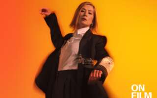 Actor Rosamund Pike in Inter Alia. She is wearing a suit and standing in front of a bright orange background.