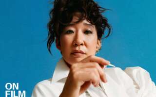 Photograph of Sandra Oh wearing a white blouse with her hair tied up against a blue background. Her hand partially covers her body.