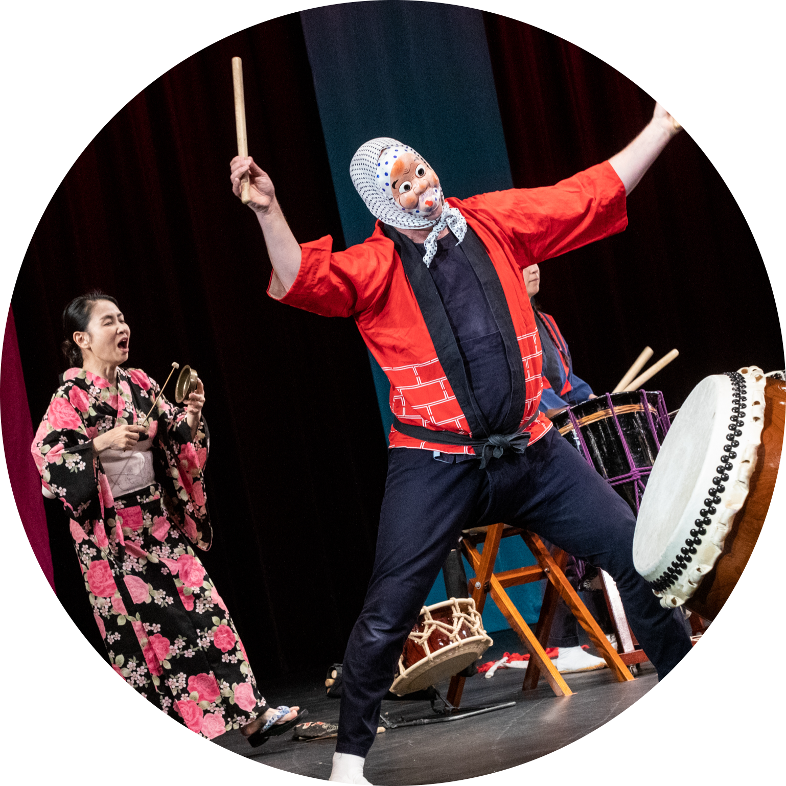 Drummer performing in a traditional Japanese Hyottoko mask holds drumsticks outstretched in the air over a large Taiko drum. A woman in a floral kimono sings and plays small instrument behind him.