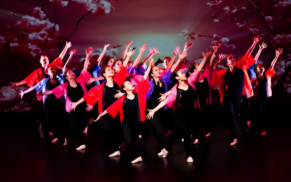 An ensemble of dancers strike a pose on stage. They wear black jumpsuits with red overcoats, and are lit by stage lights from above.