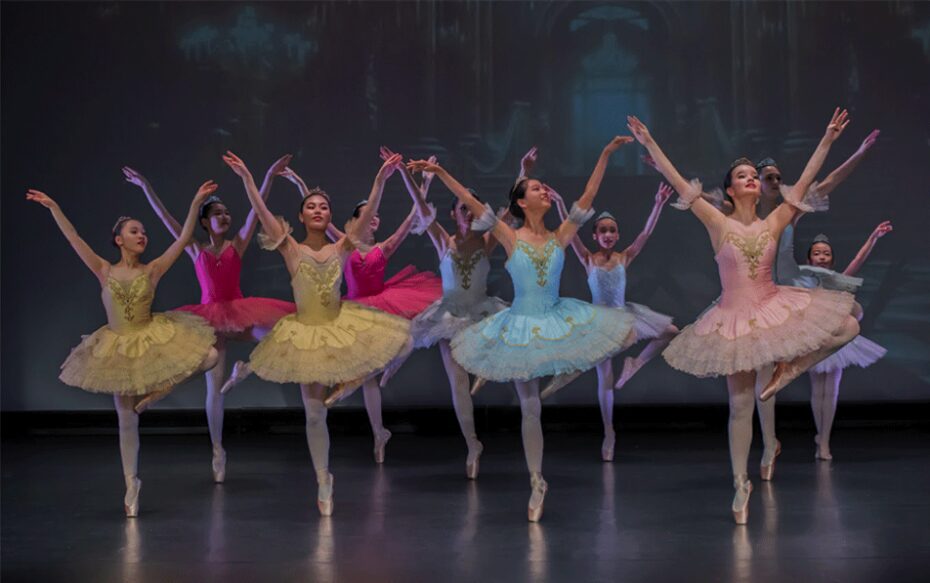 Nine ballet students strike a pose on stage, lit by stage lights. They wear yellow, blue, and pink tutus.