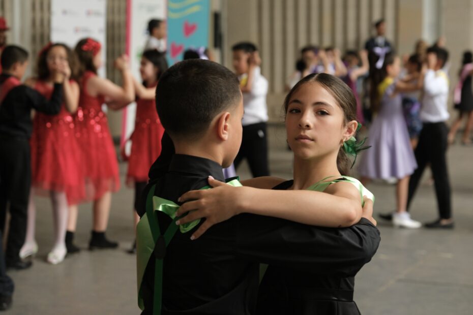 A close up photo of a pair of students dancers. They wear black. There are other students posing in the background, wearing red and purple outfits.