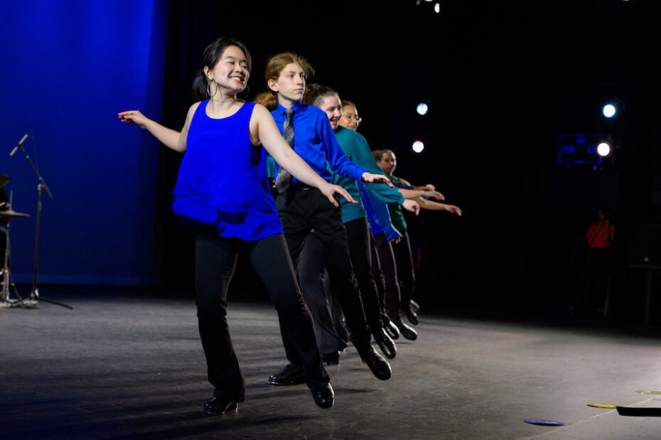 A line of young tap dancers angle away from the camera. They are lit by stage lights and are wearing blue shirts.
