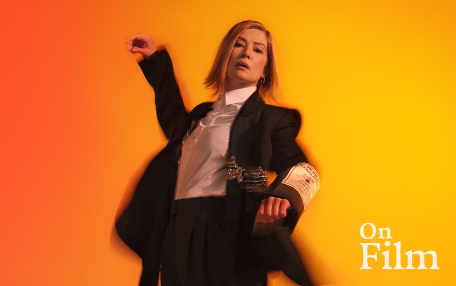 Actor Rosamund Pike in Inter Alia. She is wearing a suit and standing in front of a bright orange background.