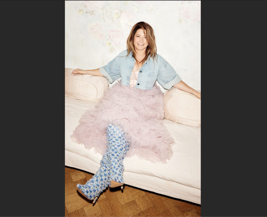 Pianist Helen Jane Long sits with her legs crossed on a white couch. She is wearing a denim coverall suit with pink tutu and smiling at the camera.