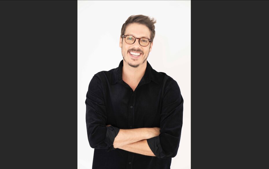 A portrait shot of Fábio Porchat over a white background. He is wearing a black sweater and glasses, smiling with his arms folded over his chest.