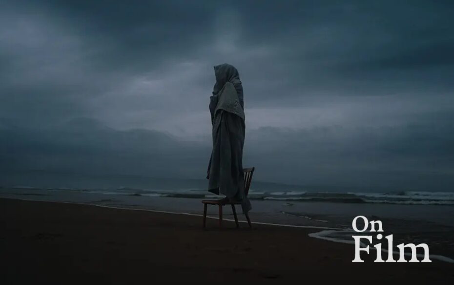 A person wrapped in a cloak stands on a chair by the sea.