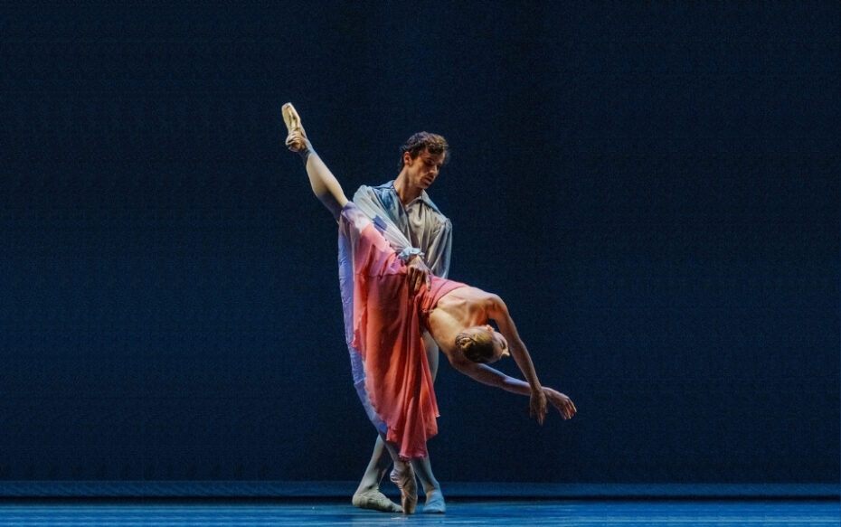 A pair of ballet dancers on stage, one dips the other towards the floor as she has her leg extended upward.. They are against a black curtain with blue lights.