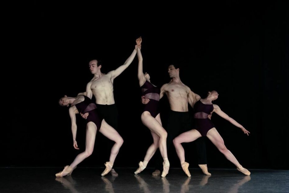 A group of modern dancers pose on stage against a black curtain. They all wear black.