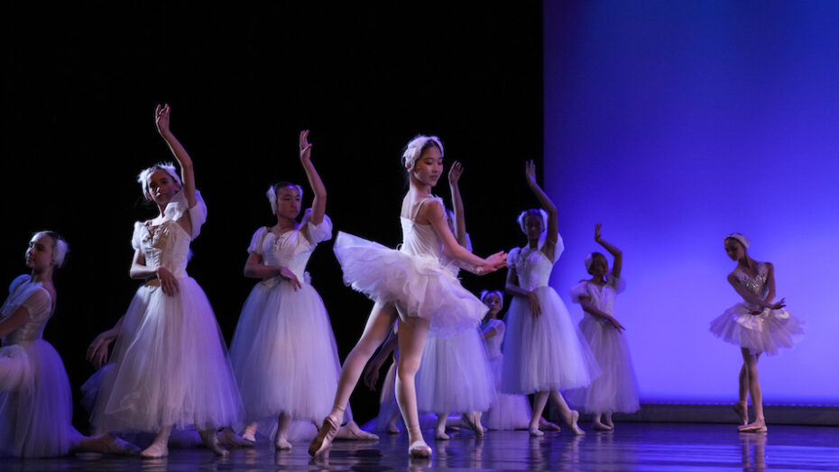 A group of ballet dancers in white tutus pose mid-dance in front of a purple lit backdrop.
