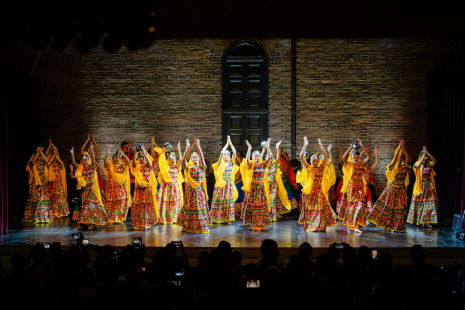 A large group of dancers on stage wearing yellow dresses. The pose with arms above their heads.