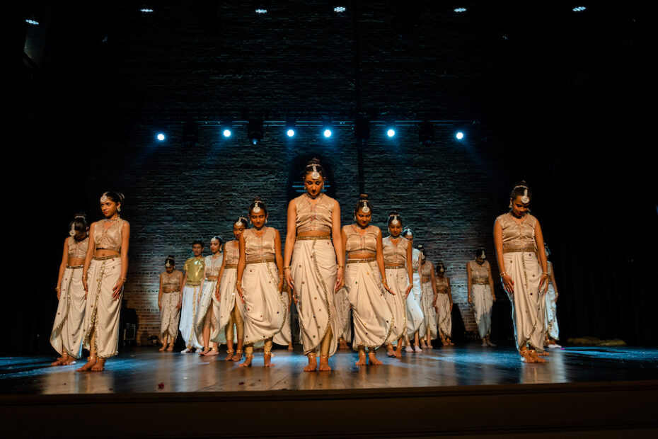 Dancers are backlit onstage wearing traditional Indian dance costumes. They all stand with their arms to their sides.
