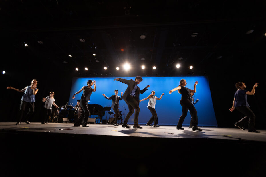 A line of dimly lit dancers tap on a full stage. The curtains are backlit in blue.