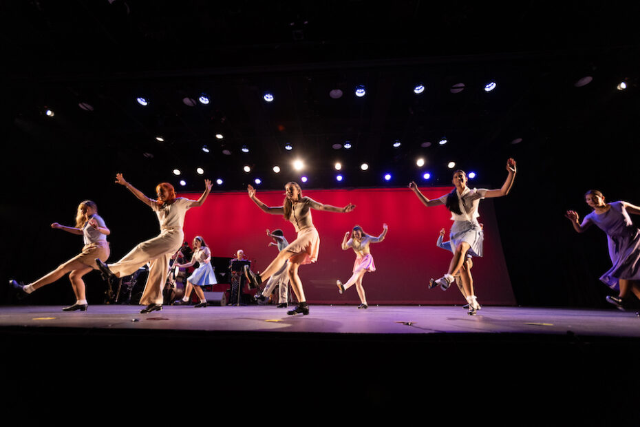 Two spaced out lines of dancers tap on a fully lit stage. A piano and jazz quartet are behind them.