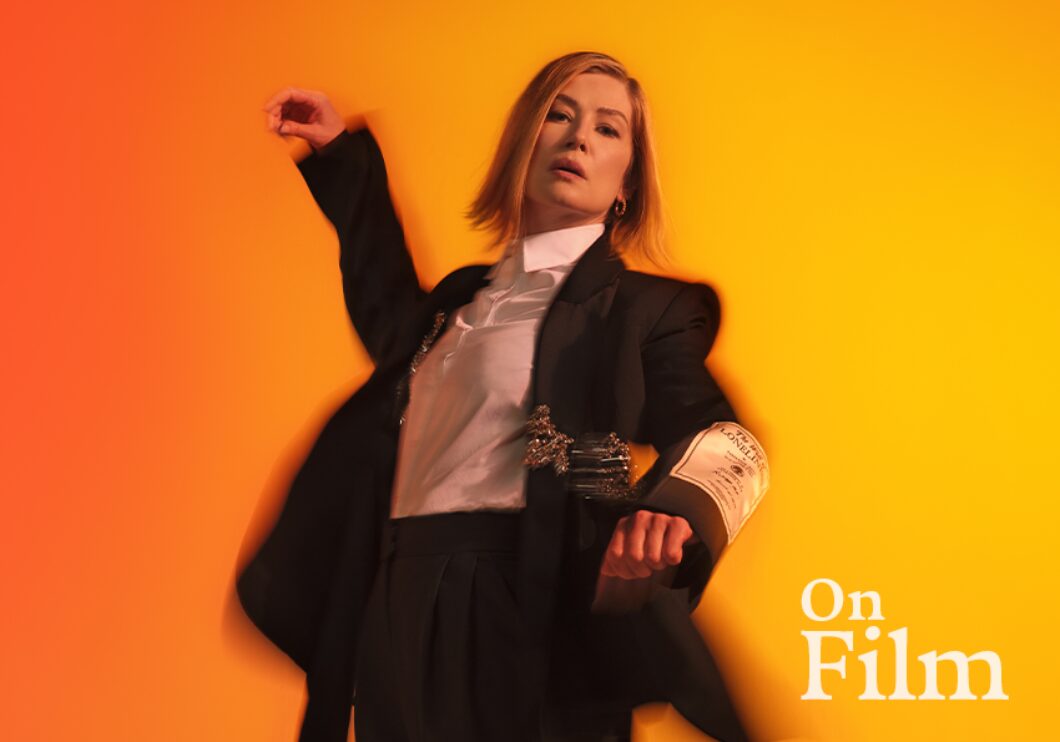 Actor Rosamund Pike in Inter Alia. She is wearing a suit and standing in front of a bright orange background.