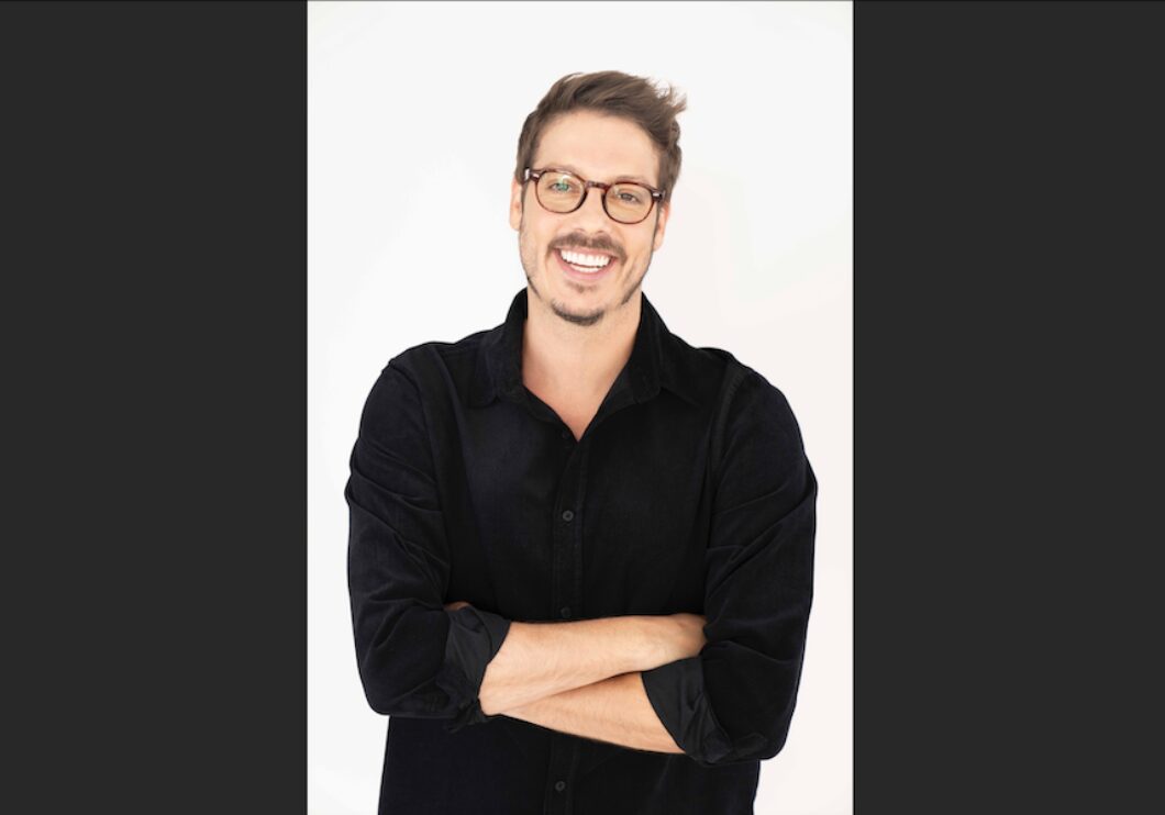 A portrait shot of Fábio Porchat over a white background. He is wearing a black sweater and glasses, smiling with his arms folded over his chest.