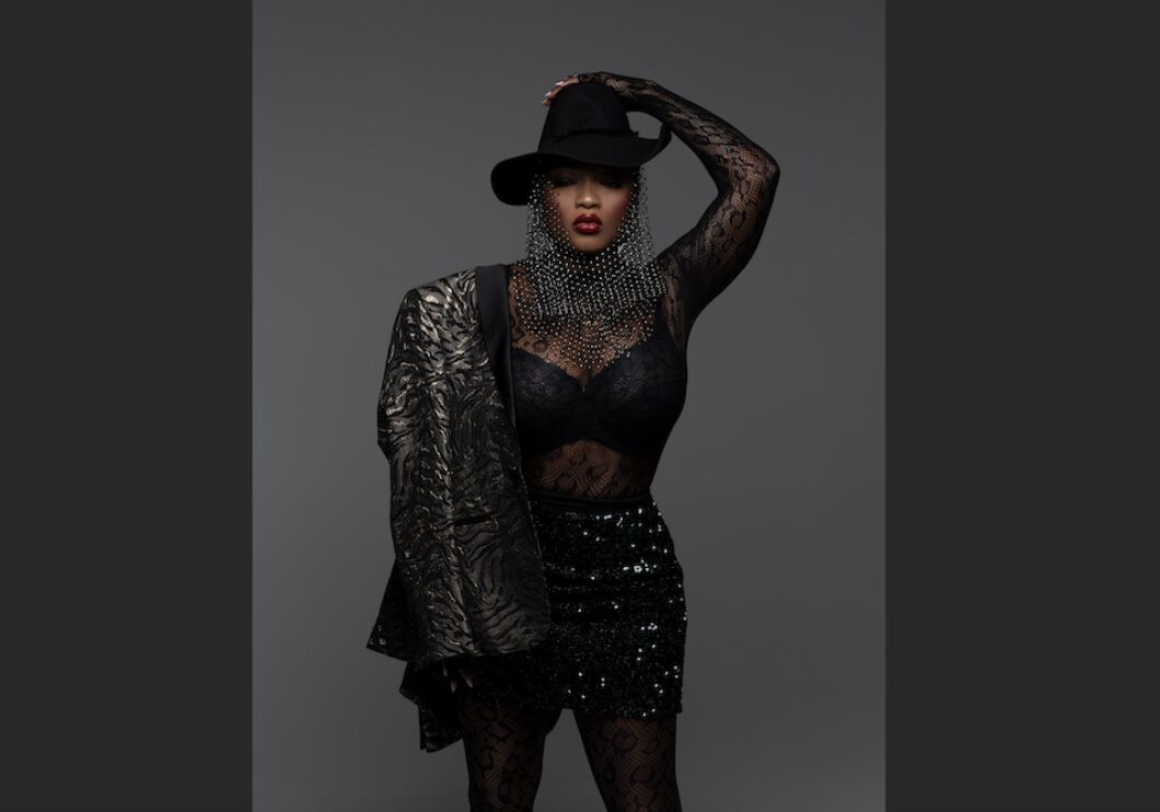 Performer Shan'L poses in an all black outfit and hat against a gray background, her face in shadow.
