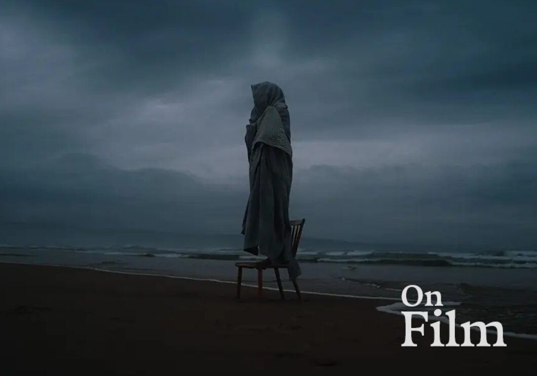 A person wrapped in a cloak stands on a chair by the sea.