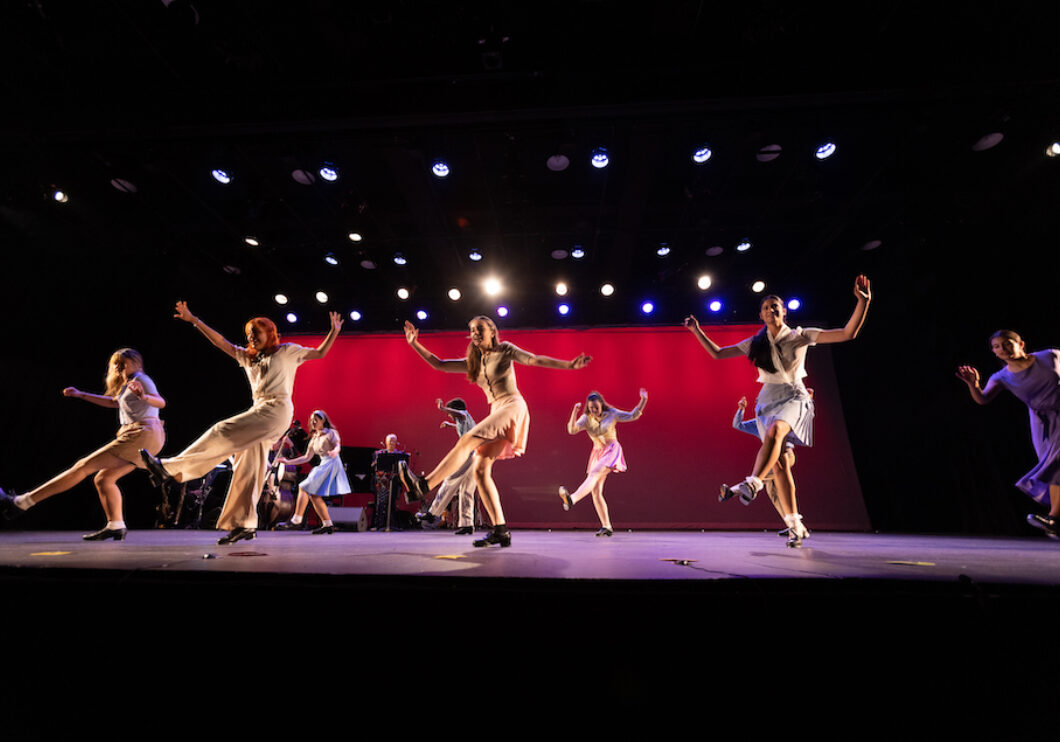 Two spaced out lines of dancers tap on a fully lit stage. A piano and jazz quartet are behind them.