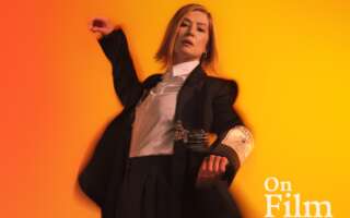 Actor Rosamund Pike in Inter Alia. She is wearing a suit and standing in front of a bright orange background.