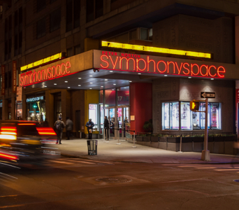 Street view of the corner of Symphony Space by night with neon lights on the marquee.