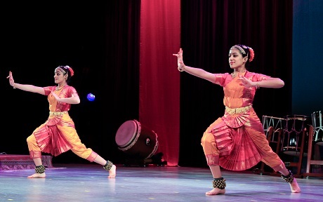 two dancers wearing colorful, flowing costumes perform a synchronized arm gesture from opposite sides of the stage