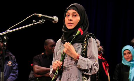 A young woman wearing a hijab stands on stage speaking into a microphone.
