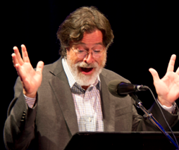 Stephen Colbert with beard standing at a mic with hands expressively raised