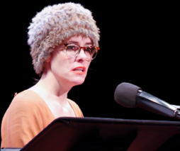 Parker Posey in hat and glasses reads on the stage.